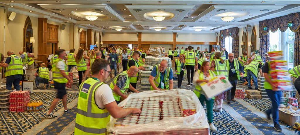 "Large group of people in a spacious, well-lit conference room, all wearing high-visibility vests branded with 'Bunzl.' The room is filled with activity as participants move quickly, stacking and organizing food and beverage items on large pallets. This is a food bank charity activity. 